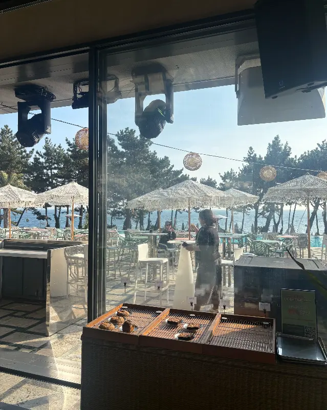 카페 내부에서 통유리창 너머로 보이는 야외 테이블과 파라솔, 바다 풍경