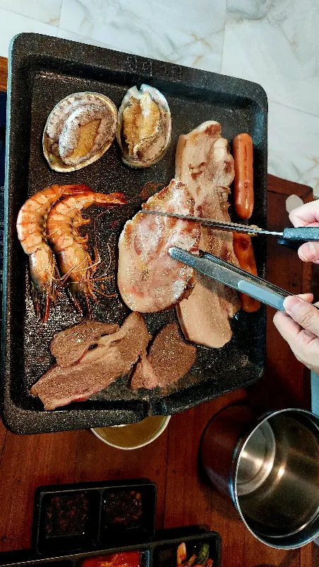 불판 위에서 맛있게 구워지고 있는 흑돼지, 전복, 새우, 소시지