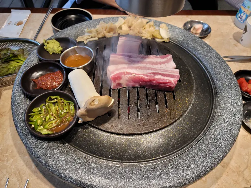 잘 구워진 삼겹살과 버섯, 마늘이 불판 위에 놓여 있고, 쌈 채소와 소스들이 주변을 둘러싸고 있다.