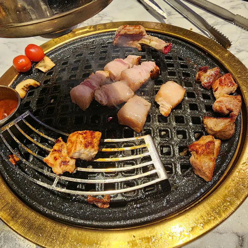 돈백꽃의 숯불 구이 모습. 직원분이 직접 고기를 굽고 있으며, 불판 위에는 흑돼지 오겹살이 노릇하게 구워지고 있다.