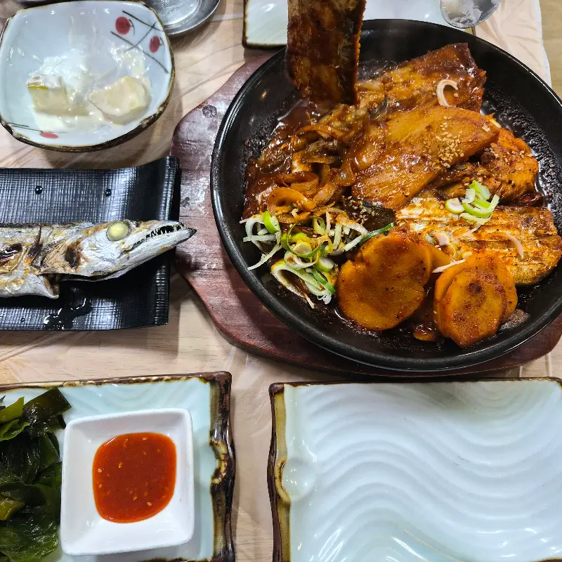 갈치구이가 검정색 긴 접시에 담겨 있고, 그 옆으로 깻잎 장아찌, 김치 등의 반찬이 놓여 있다.