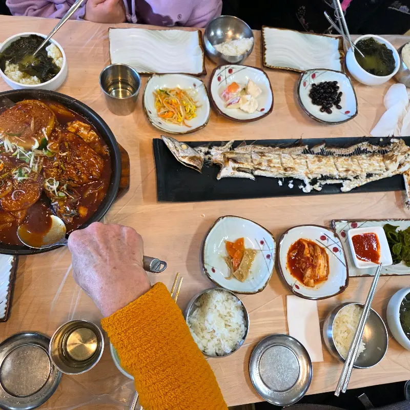 갈치조림 뚝배기를 중심으로 깻잎 장아찌, 김치, 콩나물 무침 등 다양한 반찬들이 테이블 위에 놓여 있다.