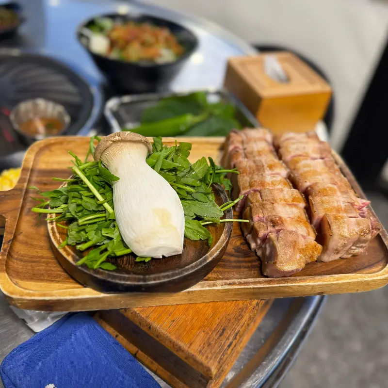 초벌된 흑돼지 근고기와 왕새송이버섯, 미나리가 나무 도마에 담겨져 있다.