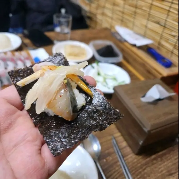 고등어회, 밥, 묵은지, 양파, 특제 소스를 김에 싸서 손으로 들고 있는 모습. 한 입에 먹기 좋은 크기로 만들어져 있다.