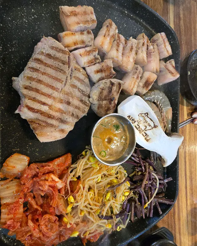 불판 위에 가지런히 놓인 흑돼지 오겹살과 목살, 김치, 콩나물, 고사리, 새우, 그리고 가운데 놓인 소스