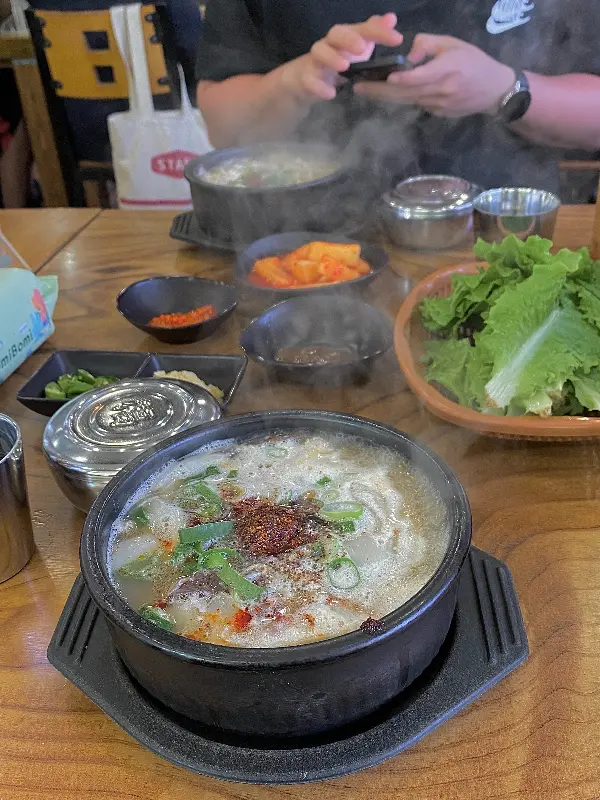 김치찌개가 뚝배기에 담겨져 김치, 쌈채소와 함께 테이블 위에 놓여있는 모습