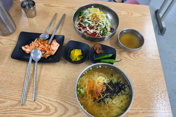 테이블 세팅 사진: 국수 한 그릇, 비빔밥 한 그릇, 김치, 깍두기, 고추, 쌈장 등이 준비되어 있다.