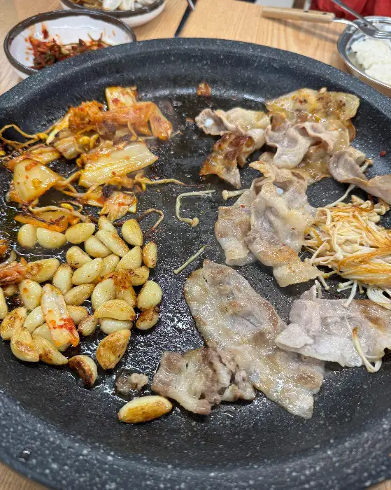 지글지글 익어가는 삼겹살과 함께 콩나물, 마늘, 김치 등이 놓인 불판 모습