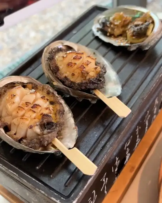 미니 화로에 두 개의 전복 구이가 꼬치에 꽂혀 구워지고 있는 모습. 겉면이 노릇하게 익어 맛있는 냄새를 풍길 것 같다.