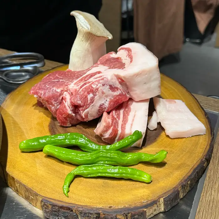신선한 흑돼지 오겹살과 목살이 나무 도마 위에 놓여 있고, 팽이버섯과 고추가 곁들여져 있습니다.