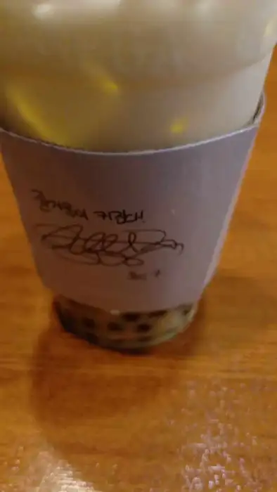 연한 녹색의 음료가 담긴 컵에 펄이 보이도록 클로즈업된 모습. 컵 홀더에는 손글씨가 적혀 있다.