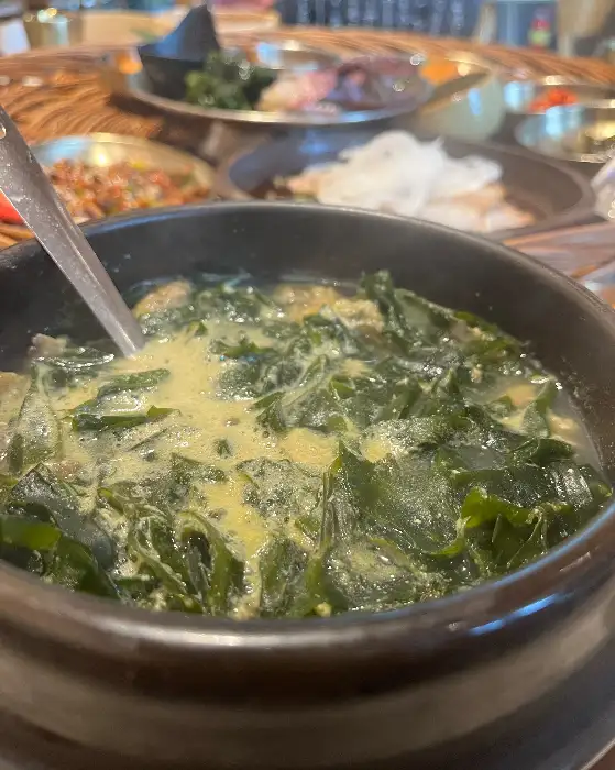 검은색 뚝배기에 푸짐하게 담긴 전복 미역국. 숟가락이 담겨 있으며, 맑은 국물 위로 전복과 미역이 보인다.