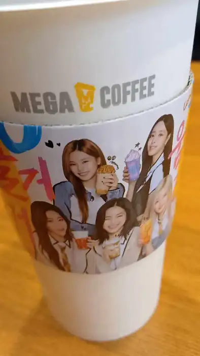 걸그룹 멤버들이 음료와 함께 포즈를 취하고 있는 컵홀더가 씌워진 음료 컵. 'MEGA COFFEE' 로고가 보인다.