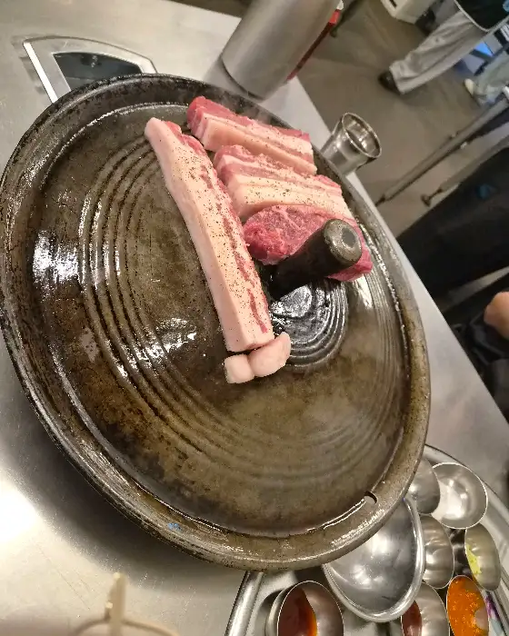 솥뚜껑 위에 가지런히 놓인 고기, 갓김치, 콩나물, 미나리
