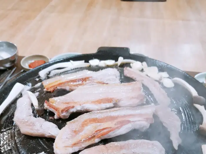 두툼한 돼지고기가 불판 위에서 익고 있으며, 주변에는 양파와 버섯이 함께 놓여 있습니다.