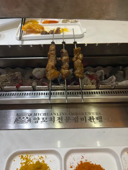 숯불 위에서 익어가고 있는 양꼬치 꼬치와 곁들임 소스들