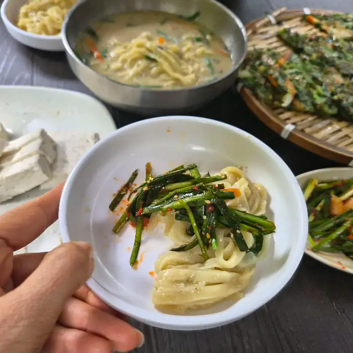 한 사람이 젓가락으로 면을 집어 올리고 있는 모습으로, 그릇 안에는 국수와 파, 당근 등 채소가 보입니다.