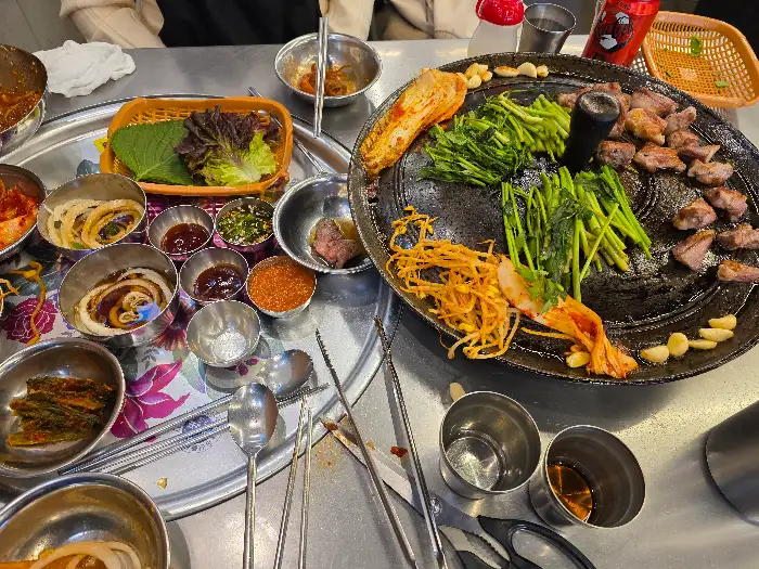 노릇하게 구워지는 삼겹살과 신선한 미나리, 그리고 다양한 곁들임 반찬이 푸짐하게 차려진 모습