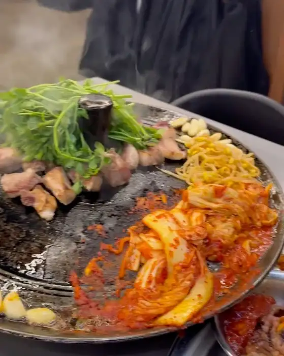 불판 위에서 익어가는 삼겹살 조각들과 신선한 미나리, 그리고 콩나물 무침과 김치가 함께 조리되는 모습