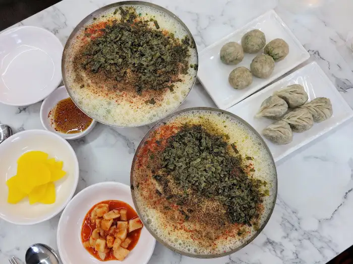 테이블 위에 칼국수 두 그릇과 굴림만두, 찐만두가 놓여 있습니다. 그 옆으로는 단무지와 김치, 그리고 양념장이 준비되어 있습니다.