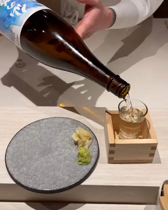 사케 병에서 잔으로 술이 따라지고 있다. 옆에는 핑크색 그릇에 생강 절임과 와사비가 놓여 있다.