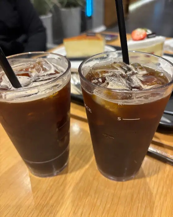  테이블 위에 놓인 두 잔의 아이스 아메리카노, 배경에는 케이크가 보인다.