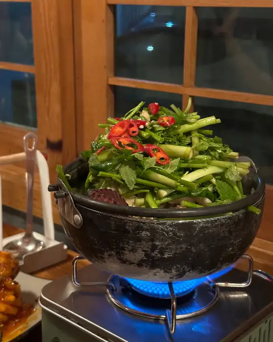 냄비 안에 푸릇푸릇한 채소가 가득 담겨 있고, 빨간 고추가 포인트로 올라가 있습니다. 가스레인지 위에 올려져 있어 막 끓기 시작하는 모습입니다.