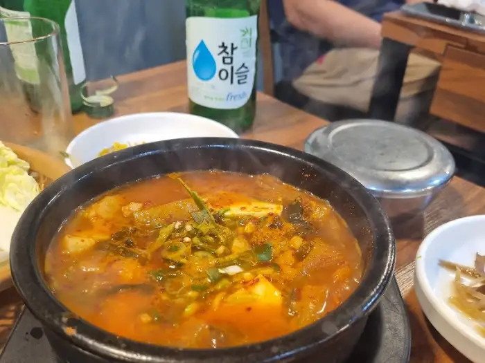 김치찌개가 뚝배기에 담겨 나오고, 그 옆에는 소주 병과 물컵이 놓여 있다.
