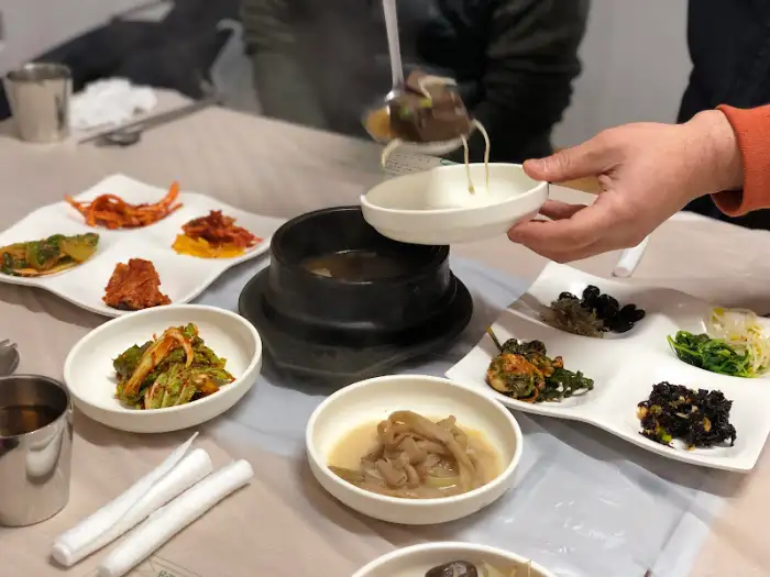 테이블 위 다양한 반찬들이 놓여 있고, 한 손으로 국물이 담긴 작은 그릇을 들고 있습니다. 여러 가지 음식을 맛보는 듯한 모습입니다.
