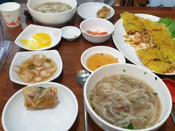 테이블에 차려진 다양한 베트남 음식들. 쌀국수, 반세오, 곁들임 메뉴가 푸짐하게 준비되어 있습니다.