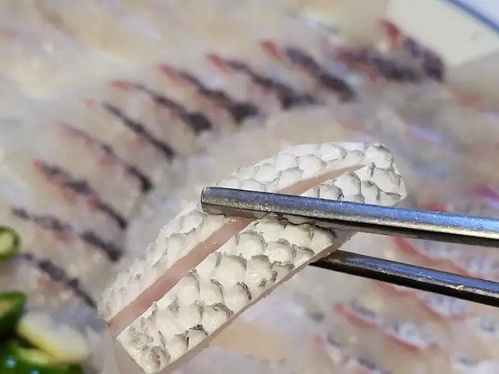 집게로 신선한 회 조각을 집어 올린 모습. 껍질 부분의 비늘 무늬가 섬세하게 표현되어 있다.