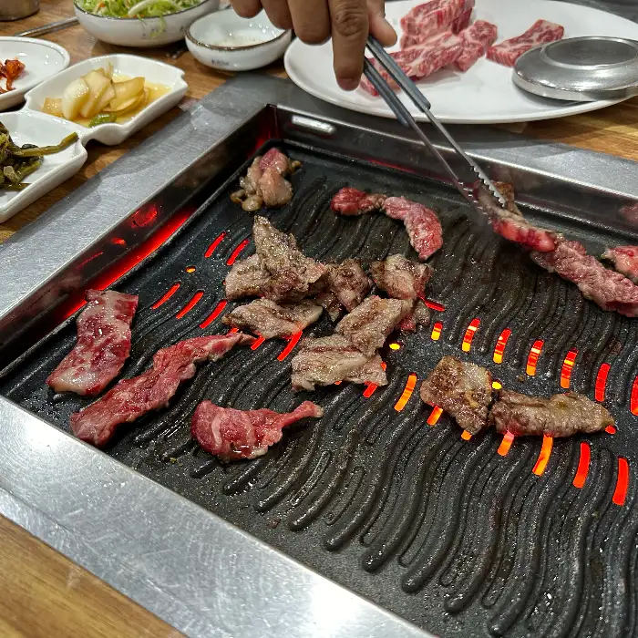 테이블 위 불판에서 약소고기가 맛있게 구워지고 있습니다. 집게로 고기를 뒤집으며 굽는 모습이 생생합니다.