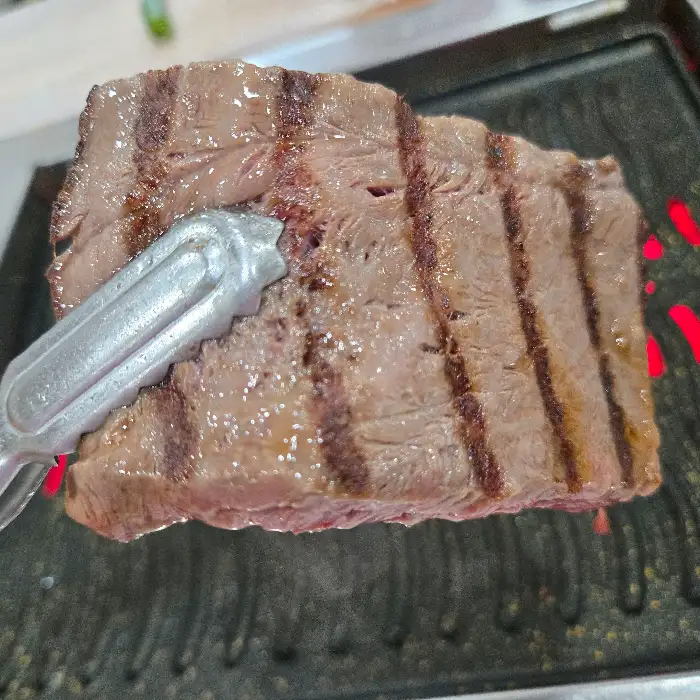 집게로 약소고기 한 점을 불판에서 집어 올리고 있습니다. 잘 익은 고기에 줄무늬가 선명합니다.