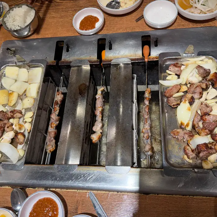 회전하며 익어가고 있는 오리 꼬치와 곁들임 채소, 밥, 소스 등이 놓인 테이블 풍경