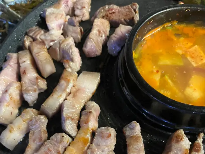 솥뚜껑 불판 위에 구워지고 있는 삼겹살과 옆에 놓인 김치찌개