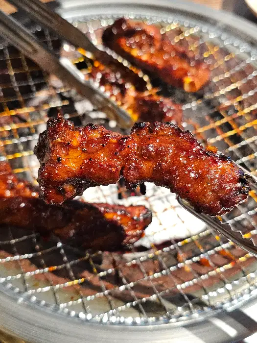 집게로 집힌 먹음직스러운 쪽갈비. 숯불 위에서 노릇하게 구워져 윤기가 흐르고 양념이 골고루 배어 있다.