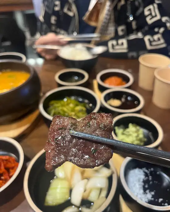 직원이 집게로 고기 한 점을 들고 있는 모습, 주변에 반찬들이 놓여있다
