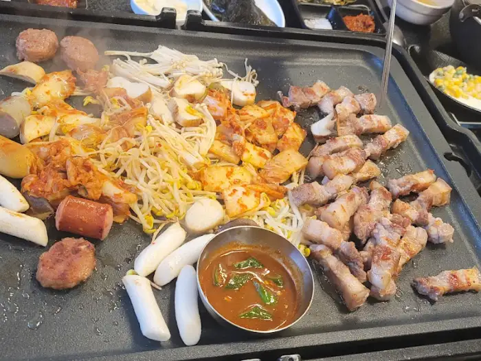 돌판 위에서 익어가는 삼겹살과 곁들임 음식들, 그리고 떡, 햄 등이 보입니다.