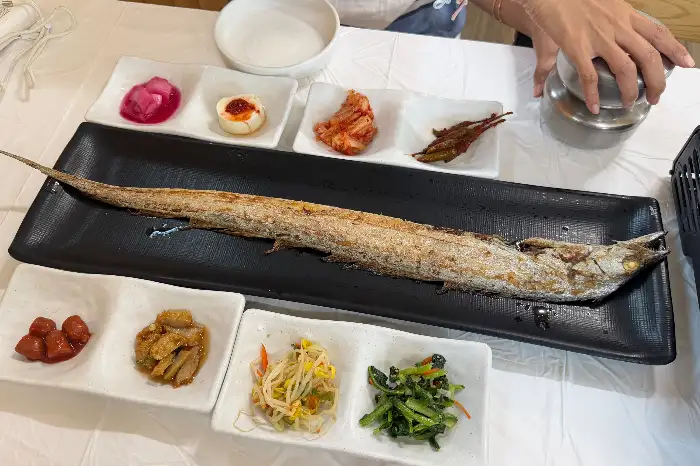 검은색 긴 접시에 통갈치구이가 놓여 있고, 주변에 작은 접시들에 김치, 장아찌 등 반찬이 담겨 있음