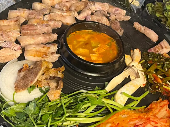 솥뚜껑 불판 위에 김치찌개, 삼겹살, 쌈 채소, 버섯 등이 놓여 있는 모습