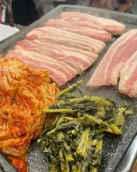 세겹집 돌판 위에서 함께 구워지는 삼겹살, 김치, 그리고 녹색 나물