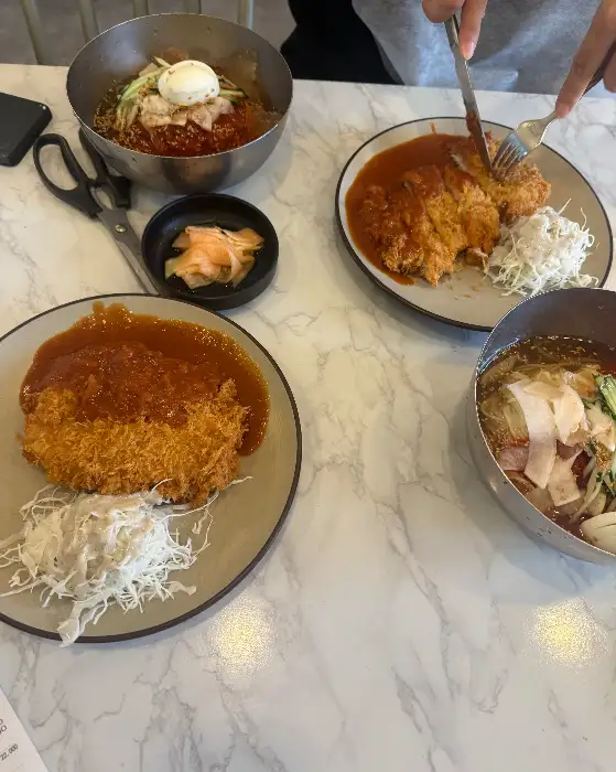 테이블 위에 밀면 두 그릇과 돈까스 두 접시가 놓여 있다. 두 가지 메뉴를 함께 즐기는 모습.